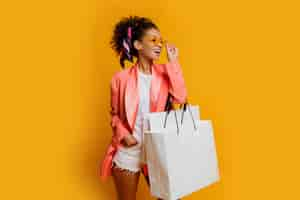 Free photo studio shot of pretty black woman with white shopping bag standing over yellow background. trendy spring fashionable look.