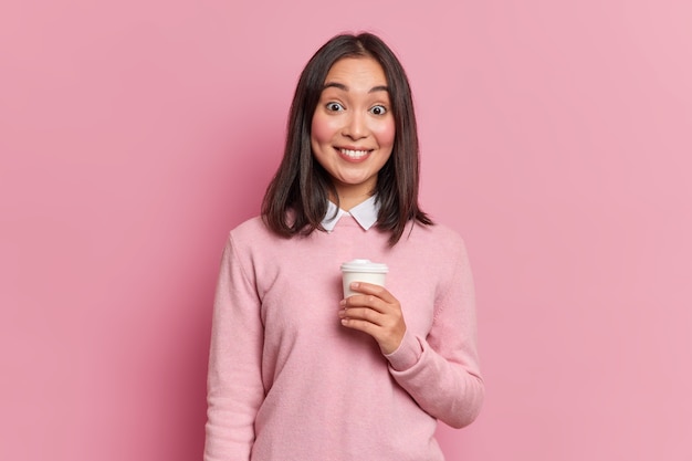 Studio shot of pretty brunette asian girl has coffee break smiles gently at camera shows white teeth dressed in casual jumper poses in studio