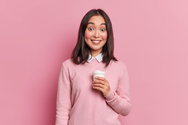 Studio shot of pretty brunette Asian girl has coffee break smiles gently at camera shows white teeth dressed in casual jumper poses in studio 
