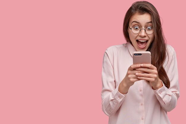 Studio shot of pretty woman stares at mobile phone, feels satisfied, stares at screen, opens mouth