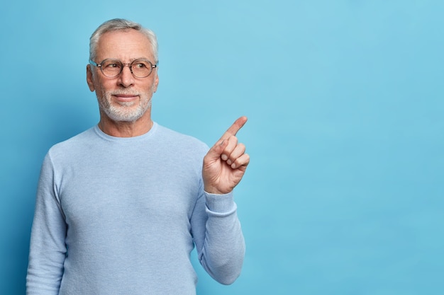 Studio shot of satisfied bearded old European man with grey hair gives recommendation suggests to use this copy space for yor advertisement dressed in casual jumper isolated on blue wall