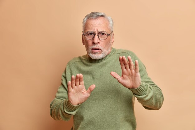 무서워하는 회색 머리 남자의 스튜디오 샷은 방어적인 제스처에 손바닥을 유지하고 공포증이 갈색 벽 위에 고립 된 캐주얼 스웨터와 안경을 착용하는 것을 본다.