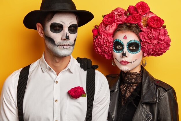 Studio shot of serious couple wears vivid makeup, celebrate traditional mexican holiday, wear wreath made of flowers, come on costume party, isolated over yellow background. Day of Death concept