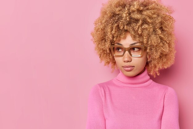 진지한 곱슬머리의 젊은 유럽 여성의 스튜디오 샷은 분홍색 배경의 빈 카피 공간에서 격리된 캐주얼 옷을 입은 무언가를 집중적인 표정으로 바라보고 있습니다.
