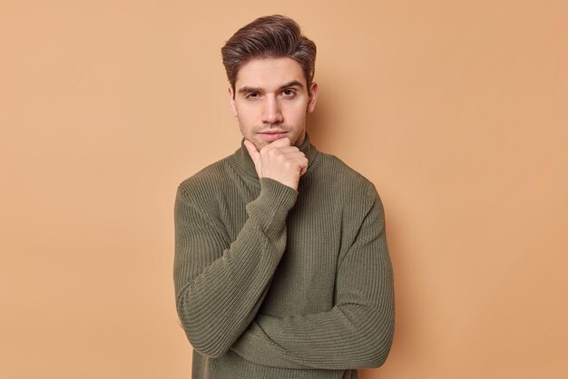 Studio shot of serious handsome adult Euroopean man holds chin focused at camera listens carefully curious plan stands determined and assertive wears casual sweater isolated over brown background.