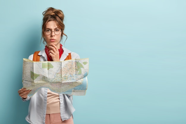 Studio shot of serious thoughtful female tourist holds chin, plans her trip, holds paper map