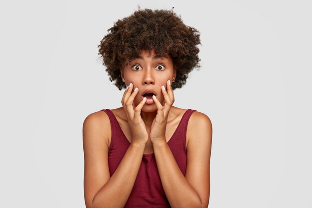 Studio shot of shocked African American girl with surprised expression, stares with bugged eyes, keeps mouth opened