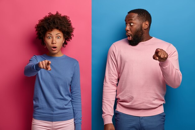 Studio shot of shocked ethnic woman and man notices very strange object, points directly at camera, cannot believe eyes