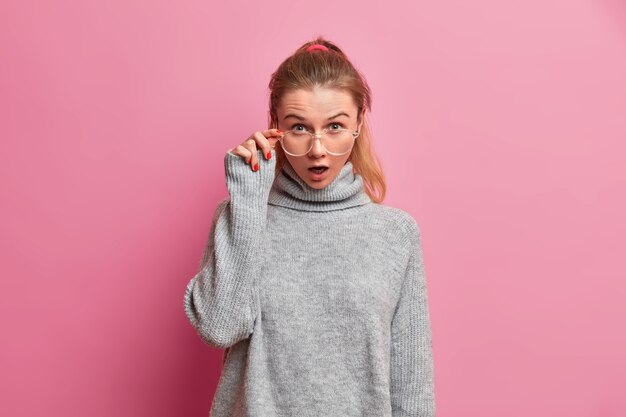 Studio shot of shocked European female model stares through transparent eyeglasses keeps mouth widely opened from astonishment