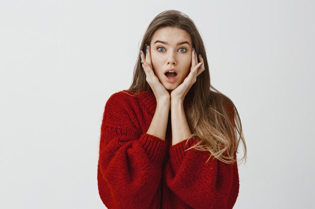 Studio shot of shocked overwhelmed attractive european girlfriend in red loose sweater, holding palms on cheeks and dropping jaw, hearing shocking news or rumor, gasping over gray wall