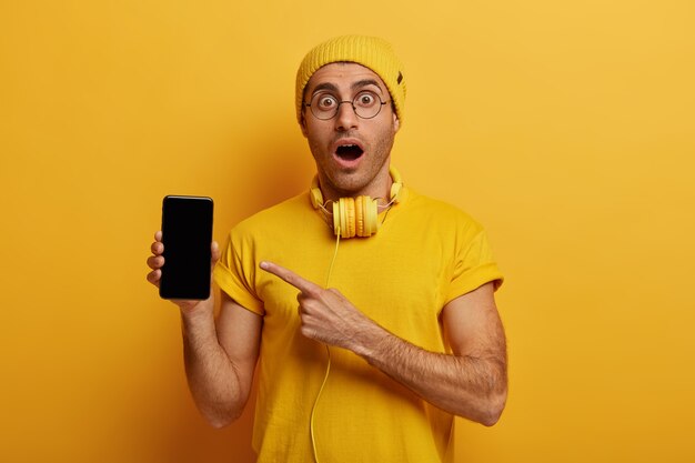 Студийный снимок потрясенного молодого человека указывает на дисплей смартфона, показывает черный экран, демонстрирует современный продукт