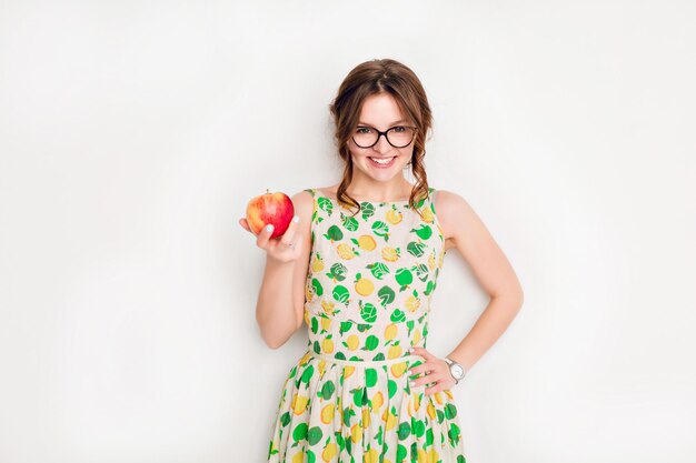 Studio shot of a smiling brunette girl smiling widely. Girl wears black glasses and yellow and green dress. She holds a red apple in her right hand.