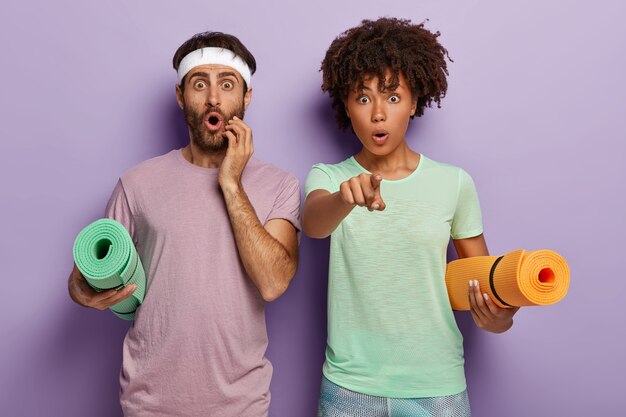 Studio shot of sporty woman and man holds yoga mats, shocked and stares straightly at camera