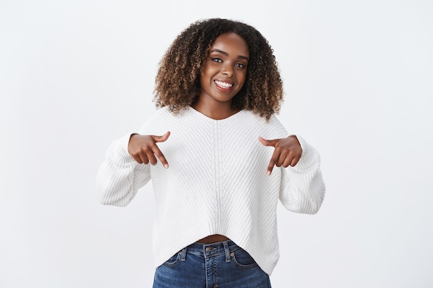 Studio shot of stylish african-american joyful female coworker in sweater pointing down and showing awesome promotion recommending look and check out advertisement, smiling happily over white wall