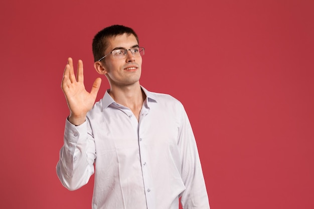 Studio shot of a stylish young person in a classic white shirt, black watches and glasses saying hello to someone while posing over a pink background. Stylish haircut. Sincere emotions concept. Copy s
