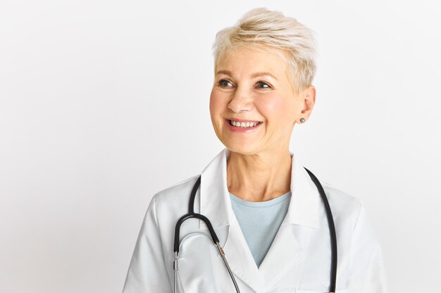 Studio shot of successful blonde middle aged female therapist posing isolated with broad happy smile wearing white medical gown and stethoscope around her neck