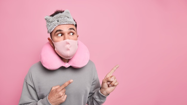 Studio shot di un uomo adulto sorpreso indossa una maschera protettiva per la maschera da notte sul cuscino da viaggio per il viso intorno al collo scioccato da qualcosa di inaspettato che punta sullo spazio