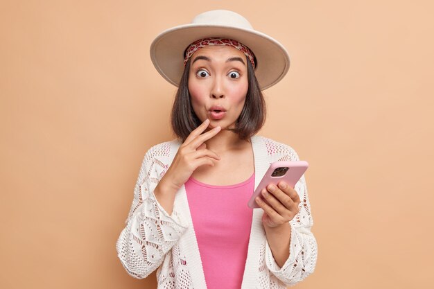 Studio shot of surprised Asian woman wears fedora stylish clothing has very shocked face gets unexpected message or comment under her post in social networks holds mobile phone uses internet