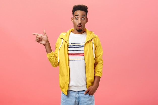 Studio shot of surprised impressed stylish african american male student in trendy yellow jacket folding lips in wow sound raising eyebrows from amazement pointing left questioned over pink wall