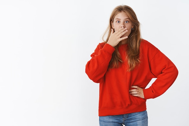 Studio shot of surprised and wondered attractive young slender good-looking woman holding hand pressed to mouth, gasping looking shocked and amazed at front, learning amusing unexpected news