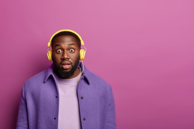 Studio shot of terrified black man stares at camera with bugged eyes, hears surprising news, wears modern stereo headphones