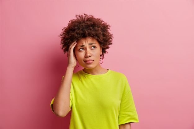 Studio shot of thoughtful puzzled curly haired woman keeps hand on head focused aside smirks face dressed in basic green t shirt isolated over pink studio wall has headache