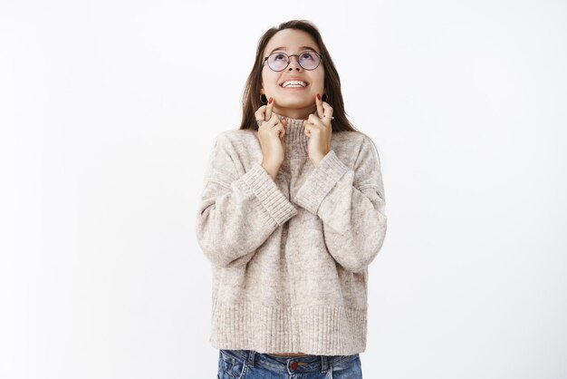 Studio shot of thrilled and happy excited attractive young woman in glasses and sweater crossing fingers for good luck looking up with broad smile praying making wish asking god for dream come true