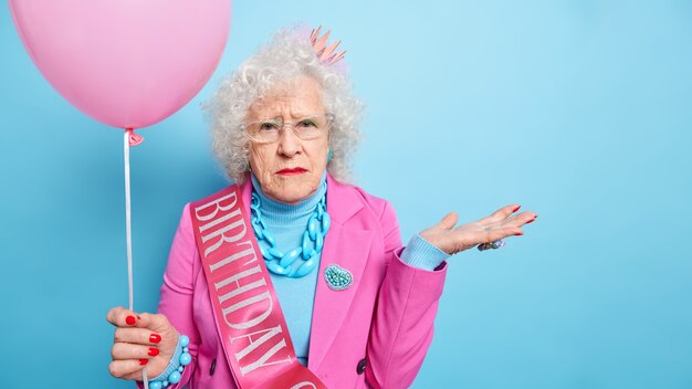 Studio shot of wrinkled grey haired elderly woman raises palms looks seriously, dressed in festive clothes holds inflated balloon celebrates birthday