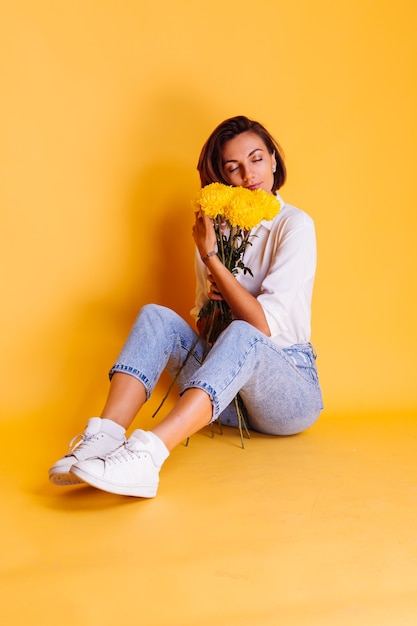 Studio girato su sfondo giallo felice indoeuropea donna capelli corti indossando abiti casual, camicia bianca e pantaloni in denim azienda bouquet di astri gialli