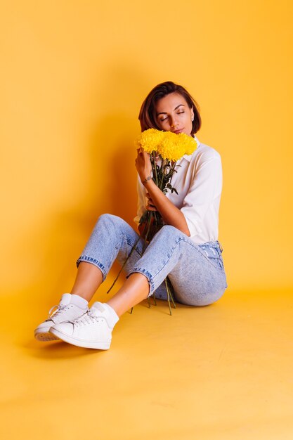 Studio shot on yellow background Happy caucasian woman short hair wearing casual clothes white shirt and denim pants holding bouquet of yellow asters