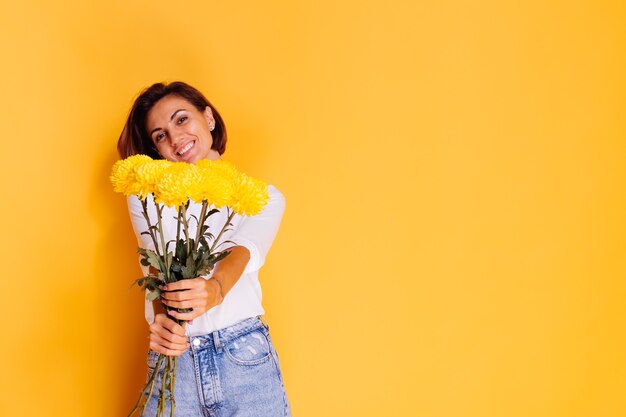 Studio shot on yellow background Happy caucasian woman short hair wearing casual clothes white shirt and denim pants holding bouquet of yellow asters