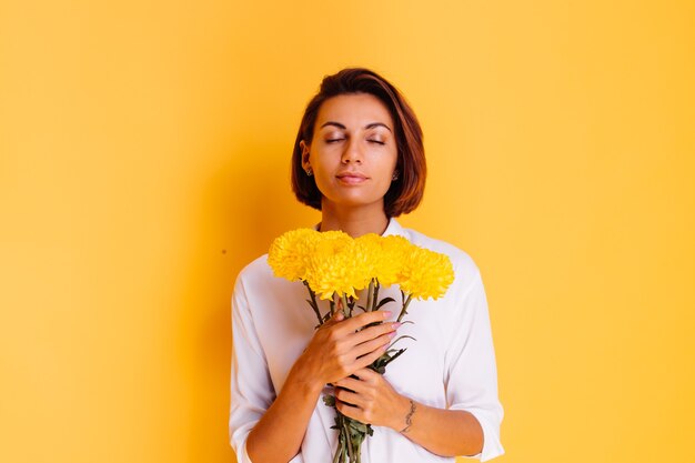 Studio shot on yellow background Happy caucasian woman short hair wearing casual clothes white shirt and denim pants holding bouquet of yellow asters