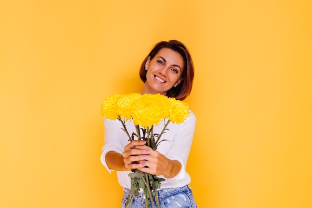 Studio shot on yellow background Happy caucasian woman short hair wearing casual clothes white shirt and denim pants holding bouquet of yellow asters