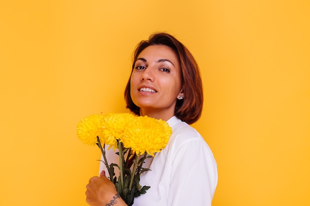 Studio shot on yellow background Happy caucasian woman short hair wearing casual clothes white shirt and denim pants holding bouquet of yellow asters