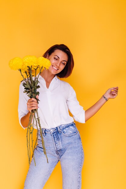 Studio shot on yellow background Happy caucasian woman short hair wearing casual clothes white shirt and denim pants holding bouquet of yellow asters