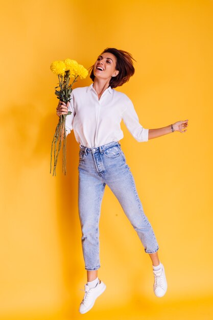 Студия выстрелил на желтом фоне Счастливая кавказская женщина с короткими волосами в повседневной одежде, белая рубашка и джинсовые брюки, держит букет желтых астр