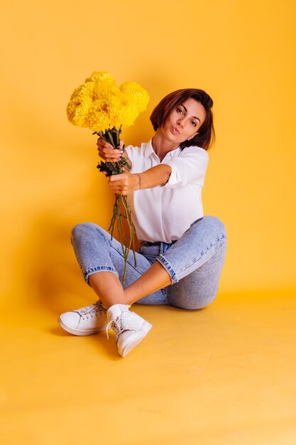 Studio shot on yellow background Happy caucasian woman short hair wearing casual clothes white shirt and denim pants holding bouquet of yellow asters