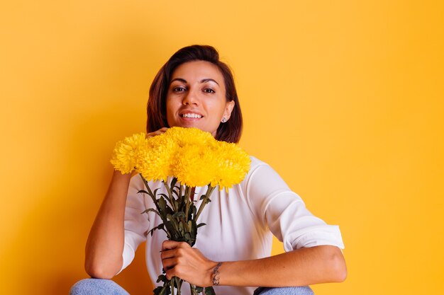 Studio shot on yellow background Happy caucasian woman short hair wearing casual clothes white shirt and denim pants holding bouquet of yellow asters