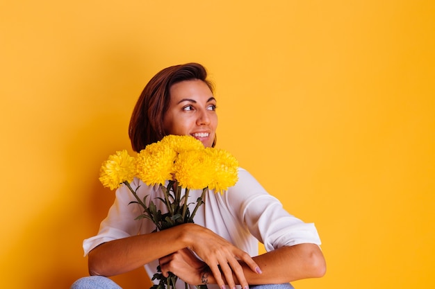 Studio shot on yellow background Happy caucasian woman short hair wearing casual clothes white shirt and denim pants holding bouquet of yellow asters