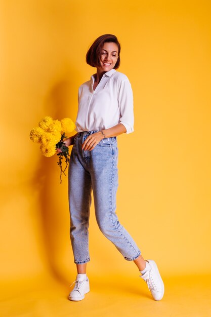 Студия выстрелил на желтом фоне Счастливая кавказская женщина с короткими волосами в повседневной одежде, белая рубашка и джинсовые брюки, держит букет желтых астр