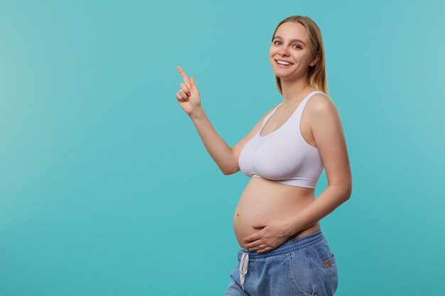 Foto gratuita studio shot di giovani piuttosto bianchi con testa futura mamma tenendo la mano sul suo ventre e mostrando allegramente verso l'alto con il dito indice, in piedi su sfondo blu