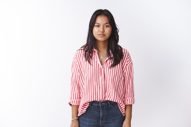 Studio shot of young vietnamese female in pink striped blouse and jeans standing relaxed as staring at camera focused, holding arms at side with casual expression posing against white wall