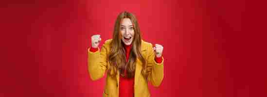 Бесплатное фото Студия што веселой возбужденной симпатичной рыжей девушки в желтом пальто, поднимающей сжатые кулаки от радости