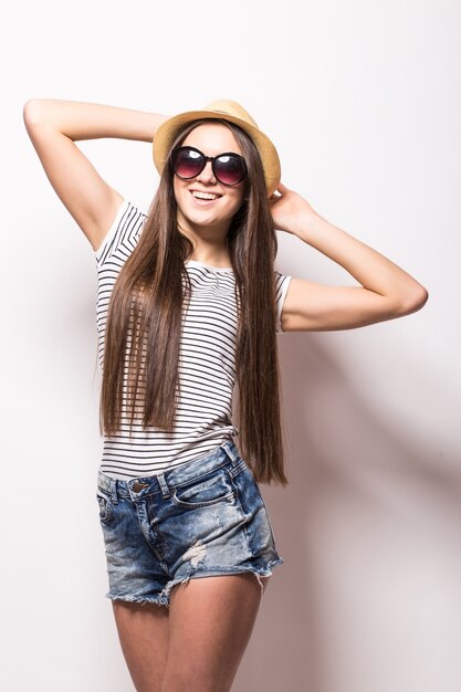 Studio view of cheerful woman with toothy smile, wears trendy sunglasses, straw hat, summer dress, isolated over white wall