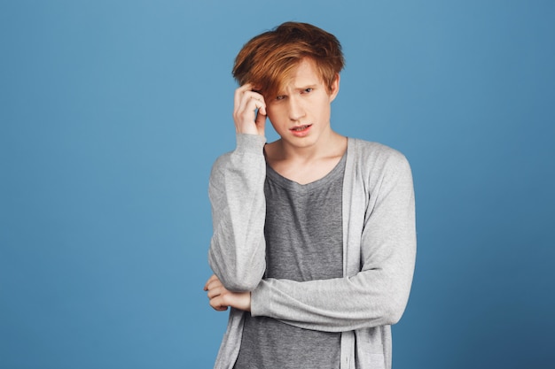 Study concept. Close up portrait of concentrated young attractive ginger student in gray clothes holding hair with hand, tying to answer teacher's question, being unsure and stressed.