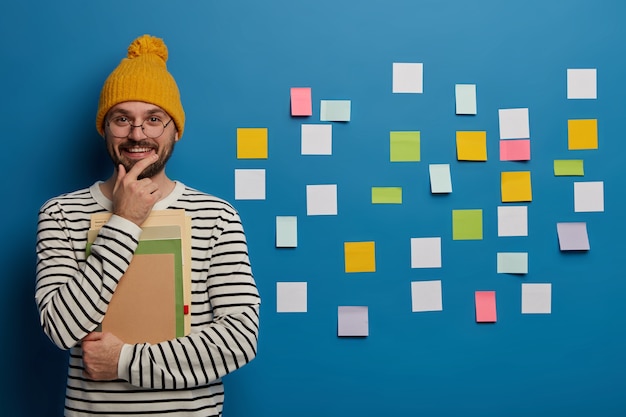 Studying, learning and education concept. Cheerful male student holds chin, smiles happily, stands with notebook and textbook, dressed in fashionable clothing, uses sticky notes on blue wall