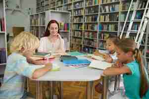 Free photo studying. young adult smiling woman with long red hair and involved school children reading and writing at table in light library