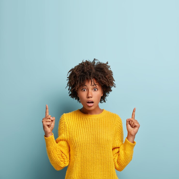Free Photo stunned attractive female points index fingers above, shows blank space for advertising content, hs bated breath, impressed by scary thing, wears yellow jumper, poses over blue wall. omg concept