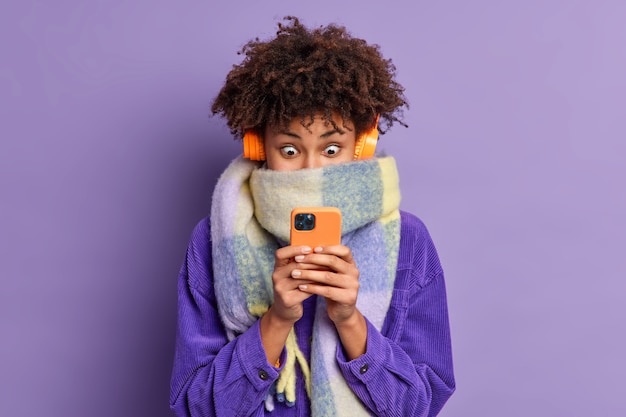 Free photo stunned beautiful millennial girl with curly bushy hair stares at smartphone display scrolls news in internet wears wireless headphones velvet jacket scarf around neck.