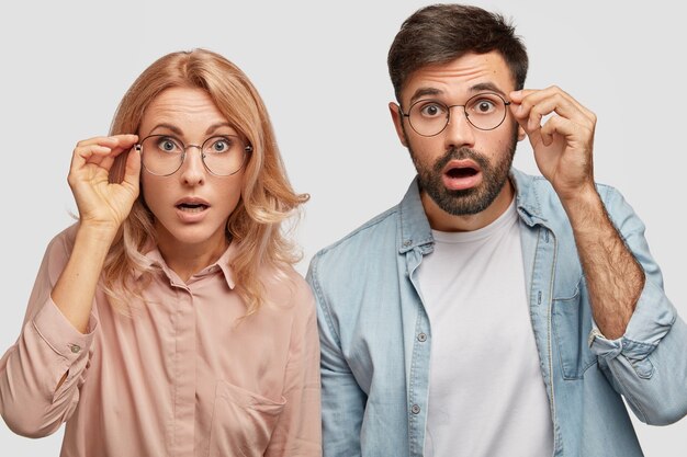 Stunned beautiful woman and his man partner, looks with eyes full of disbelief, stare through glasses